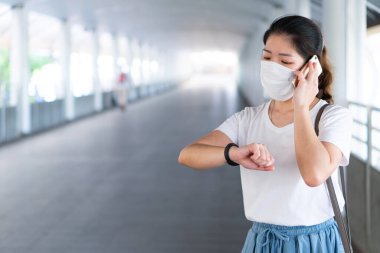 Asyalı genç bir kadın yüz maskesi takıyor, akıllı telefon kullanıyor ve Coperid-19 ya da Coronavirus salgını sırasında şehirde yürüyor. Sosyal uzaklık ve yeni normal yaşam tarzı kavramı