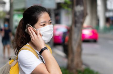 Asyalı genç bir kadın yüz maskesi takıyor, akıllı telefon kullanıyor ve Coperid-19 ya da Coronavirus salgını sırasında şehirde yürüyor. Sosyal uzaklık ve yeni normal yaşam tarzı kavramı