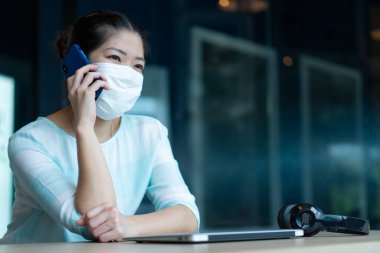 Yüz maskesi takan ve kulaklık takan Asyalı genç bir kadın. Coperid-19 ya da koronavirüs salgını sırasında evden bilgisayar kullanarak çalışıyor. Sosyal uzaklık ve yeni normal yaşam tarzı kavramı