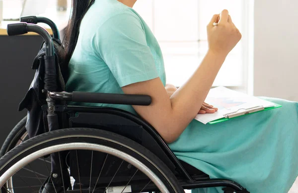young asian disabled woman with smiling face sitting in the wheelchair and using computer to discuss project with her colleagues in the working office. disability and handicapped concept