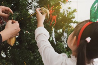 Asyalı anne ve küçük kızı Noel Baba şapkası takıyor ve Noel ağacını süslemek için süs kullanıyorlar. aile kavramı