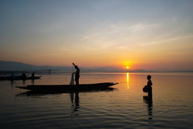 Asyalı balıkçı teknede balık tutmak için balık ağı kullanırken oğlu gün doğumunda gölde durup kavanozu tutuyor.