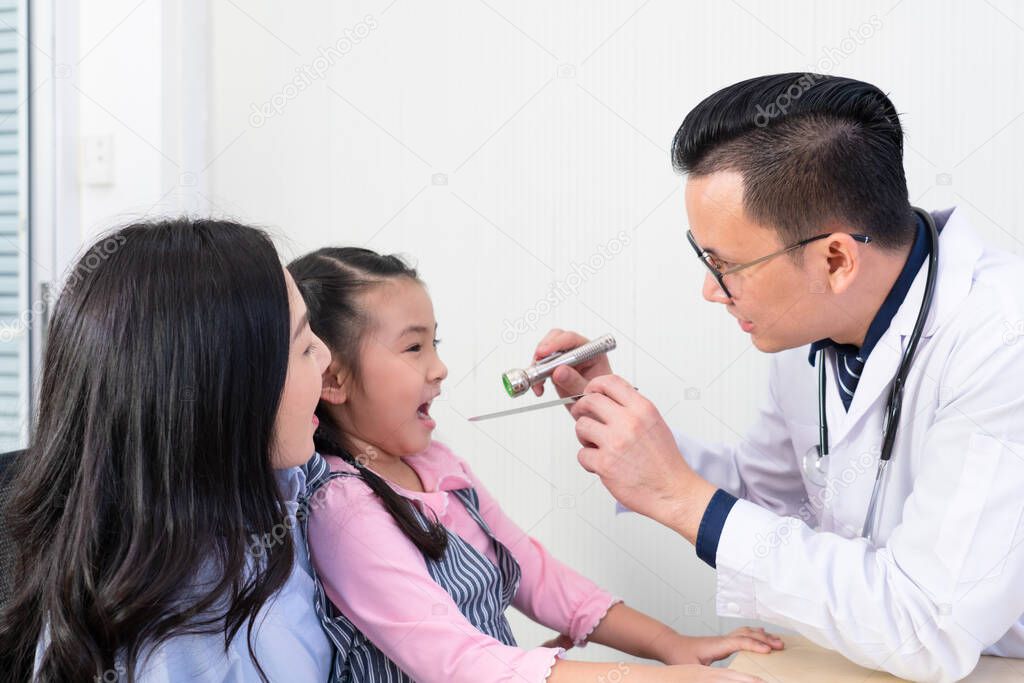 asian doctor using light torch and tongue depressor to examine young girl who has sore throat. pediatrician and healthcare concept