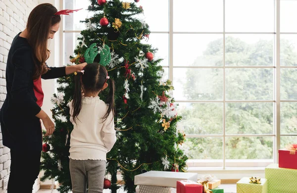 Asyalı anne ve küçük kızı Noel Baba şapkası takıyor ve Noel ağacını süslemek için süs kullanıyorlar. aile kavramı