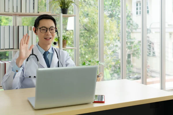 young asian doctor wearing headphone and using computer to give consult to the patient via video call in the hospital office. telemedine and healthcare concept