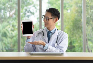 Genç Asyalı doktor hastaya danışmak için hastane ofisinde bilgisayar ve tablet kullanıyor. telemedin ve sağlık konsepti