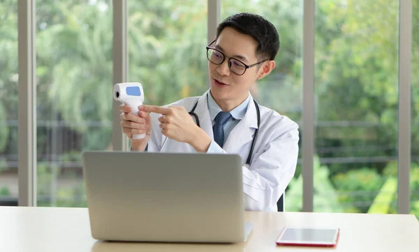 young asian doctor using computer with video call conference application to teach people how to use infrared thermometer to measure temperature . telemedicine and healthcare concept