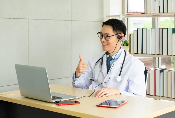 young asian doctor wearing headphone and using computer to give consult to the patient via video call in the hospital office. telemedine and healthcare concept