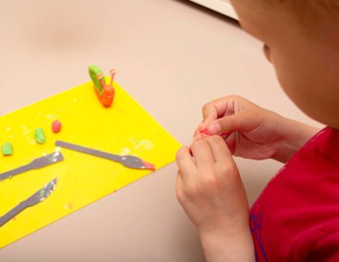 Kırmızı kazaklı bir çocuk plastikçiden salyangoz heykeli yapıyor. Masanın üzerinde sarı bir tahta ve çok renkli bir plasticin var. Coronavirüs salgını sırasında evde bir çocukla uğraştım..