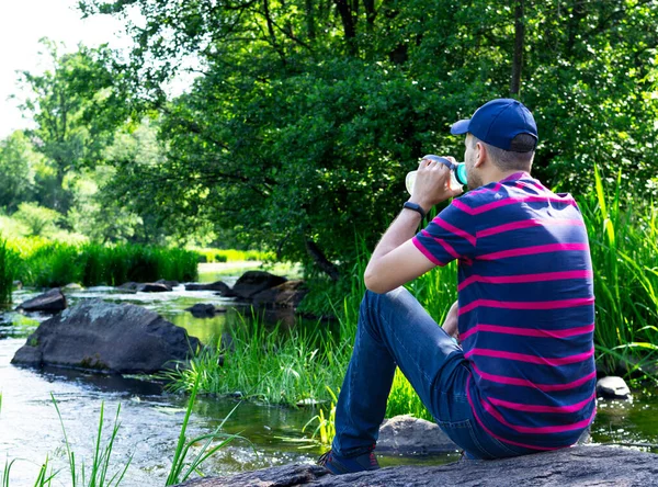 A tourist is breathing on a stone by the river. Traveler quenches thirst with water. A man sits on a stone in the forest and drinks water from a bottle