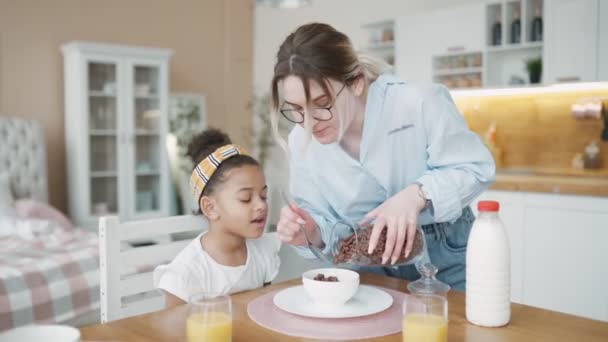 Frukost i köket: ung vacker mor häller majs flingor med mjölk, bedårande liten dotter börjar äta med glädje. Vårdande mamma förbereder flingor frukost för sin söta lilla afrikanska flicka — Stockvideo