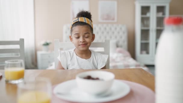 Affascinante bambina africana afroamericana con una benda gialla nei capelli mangia felicemente corn flakes con il latte. Colazione per bambini in cucina a casa prima di scuola. Buon umore dalla mattina in poi — Video Stock