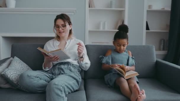 Retrato de la joven madre moderna con una hija adoptiva africana en la sala de estar. La maestra lee en el sofá, y el niño escucha con el libro en las manos. Concepto de familia feliz o educación preescolar — Vídeo de stock