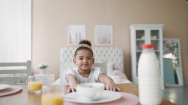Sabahları, beyaz bluzlu güzel Afrikalı kız evde, mutfaktaki masada oturur ve okuldan önce elleriyle mısır gevreği yer. Kameraya bakan çocuk portresi