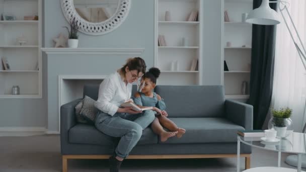 Giovane madre felice, genitore adottivo o insegnante che legge un libro alla sua piccola figlia afro-americana. Ragazza ascolta con attenzione, sorridente, seduto sul divano in soggiorno.Istruzione, insegnamento, educazione prescolare — Video Stock