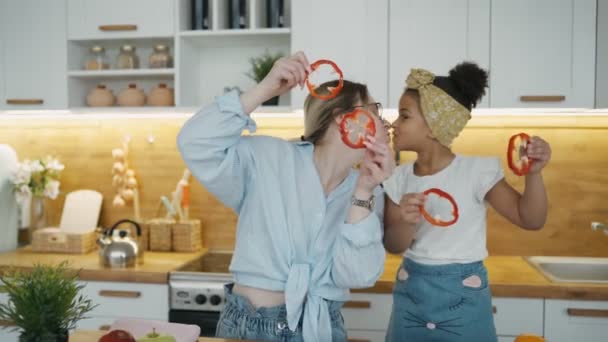 Jonge moeder en Afrikaanse dochter maken groentesalade in de keuken thuis. Familie heeft plezier samen, ogen bedekken met rode paprika cirkels die eruit zien als brillen. Lachen en kussen speelse stemming — Stockvideo