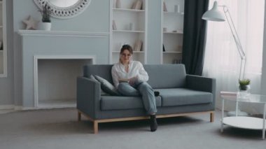 Modern oturma odasındaki pencerenin önündeki kanepede oturmuş kitap okuyan güzel, zeki bir kız. Mutlu, gülümseyen kadın öğrencinin yakın plan portresi. Evde dinlenme ve eğitim kavramı