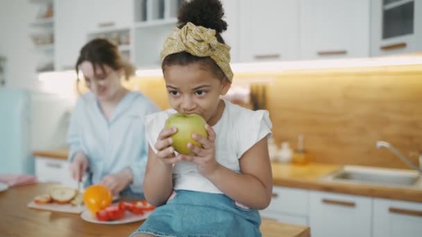 Afrikanische kleine Tochter beißt auf dem Küchentisch sitzend in grünen, saftigen Apfel. Pflegeeltern, Mütter oder Kindermädchen kochen im Hintergrund Gemüsesalat aus rotem Pfeffer. Porträt eines Mädchens mit Verband auf dem Kopf — Stockvideo