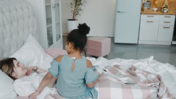 African little girl daughter in denim dress came to her mothers bedroom to wake her pinching her sister or nanny by nose. Sitting on bed in bright room with pink and white pillows and plaid in cage — Stock Video