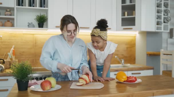 Gelukkig multiraciaal jong gezin in de keuken thuis. Internationale moeder en dochter koken lunch. Moeder snijdt een appel voor haar kleine Afrikaanse meisje.Child horloges proces hartstochtelijk en zorgvuldig — Stockvideo