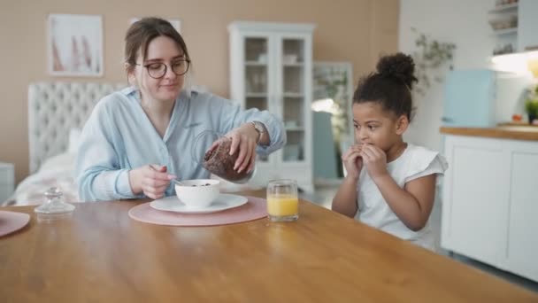 Multiraciale jonge familie zit thuis aan tafel te ontbijten in de keuken. Internationale moeder en Afrikaanse dochter eten cornflakes en drinken sinaasappelsap. Veel plezier samen voor school. — Stockvideo