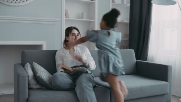 Multietnická šťastná rodina. Mladá moderní bílá matka sedí na pohovce v útulném obývacím pokoji čtení knihy. Honey africké dcera předškolního věku zábava běží k ní jako letadlo objímání a líbání — Stock video