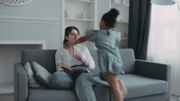 Familia feliz multiétnica. Joven madre blanca moderna sentada en el sofá en el acogedor libro de lectura de la sala de estar. Miel africana hija de preescolar diversión corre a ella como un avión abrazando y besando — Vídeo de stock