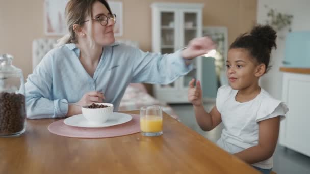 A többnemzetiségű család reggelije a modern, hangulatos konyhában. Otthon méz Afrikai óvodás lánya figyelmesen eszik kukoricapelyhet, narancslevet iszik. A lány játszik az étellel, hogy gondoskodó anya nevetni — Stock videók