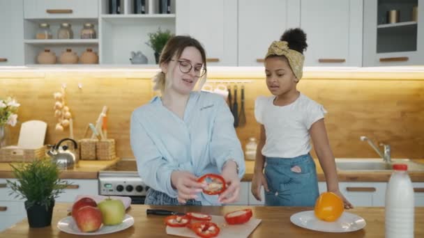 Vrolijk vrolijke moeder leert haar dochter koken, vrolijk Afrikaans meisje lacht grappige gezichten bedekken ogen met rode paprika cirkels eruit zien als brillen, verrekijker vorm. Koken in de keuken thuis — Stockvideo