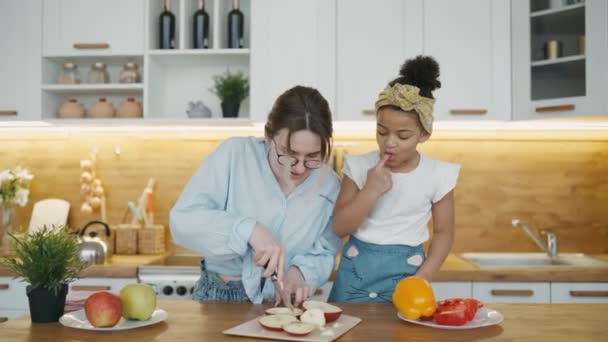 Happy interracial par nyder tid sammen hjemme, have det sjovt, mens madlavning i hyggeligt køkken. Mor skærer cirkler ud af et æble som briller til sin afrikanske datter, de laver sjov og griner – Stock-video