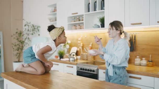 Die multiethnische Familie in der Küche des Hauses genießt ihr gesundes Müslifrühstück. Weiße junge Mutter mit Brille füttert ihre süße schwarze Tochter. Spielfreudiges Kind kaut freudig auf Tisch — Stockvideo