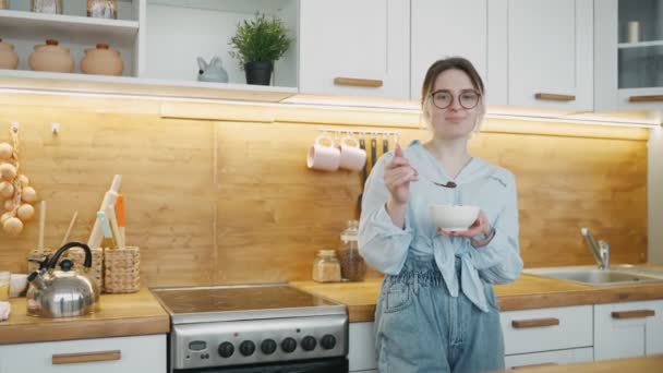 Portretul unei femei moderne fericite purtând ochelari pe fundalul bucătăriei. Fata ia micul dejun cu cereale, ţine castron şi lingură. Bucurându-se de o masă sănătoasă în picioare. Ea zâmbește uitându-se la cameră . — Videoclip de stoc