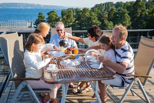 Big happy family having breakfast outdoors on terrace together, sitting around table, drinking coffee. Beautiful sea view, warm summer morning. Family bonds concept