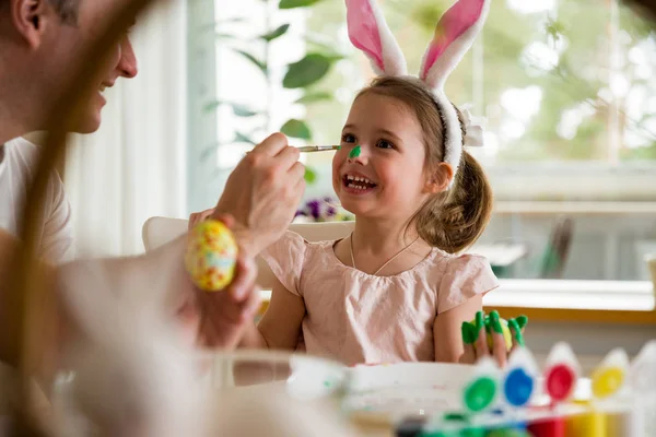 A father and daughter celebrating Easter, painting eggs with brush. Happy family smiling and laughing, drawing on face. Cute little girl in bunny ears preparing the holiday.