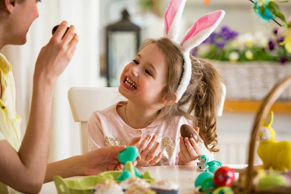 Mother and daughter celebrating Easter, eating chocolate eggs. Happy family holiday. Cute little girl with funny face in bunny ears laughing, smiling and having fun.