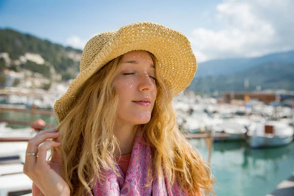 Portrait of young woman with light curly hair in straw hat enjoying sun and breeze, smiling, eyes closed. Sunny harbor with boats and yachts, green mountains on background. Enjoying life, traveling,