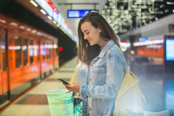Teenager girl with backpack and bike standing on metro station holding smart phone in hand, scrolling and texting, smiling and laughing. Futuristic bright subway station. Finland, Espoo