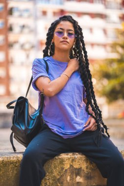 Yaşam tarzı, uzun örgülü, mor gözlükleri olan, arka planda binaları olan, parktaki bir bankta oturan esmer tenli genç kadının görüntüsü.