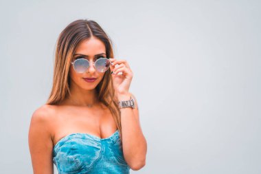 Yaşam tarzı oturumu, mavi kot elbiseli, gri arka planda güneş gözlüklü, beyaz tenli genç bir esmer.