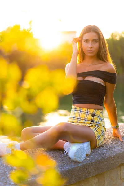 生活方式 美丽的夕阳 一个年轻的女孩坐在河边和树旁的公园里 布鲁内特 卡西亚女人 黑色紧身上衣 黄色裙子和白色运动鞋 — 图库照片
