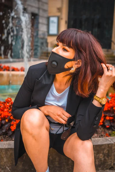 Fashion photo shoot of the new normal, a young brunette with a mask on her face from the coronavirus pandemic, sitting in the city. Lack of confidence after the covid-19, social distance