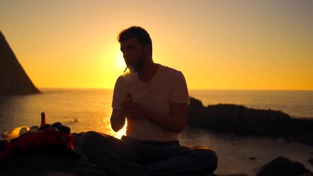 ライフスタイル 日没の灯台の海の近くの岩の上に座っての横にあるサングラスの若い男 太陽を背に日没時にタバコを灯す — ストック動画