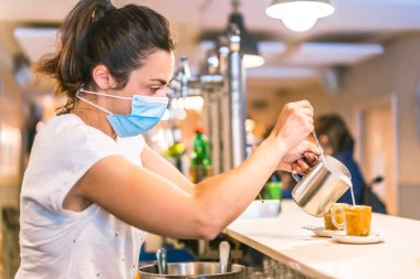Barda maske takan bir garsonla fotoğraf çekimi. Yeni normallik, koronavirüs salgınından sonra alınan güvenlik önlemleri, sosyal mesafe, covid-19. Bir fincan kahveye süt koyuyorum.