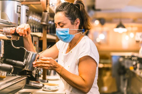 Barda maske takan bir garson. Yeni normallik, koronavirüs salgınından sonra alınan güvenlik önlemleri, sosyal mesafe, covid-19. Kahve makinesiyle sütlü kahve hazırlıyorum.