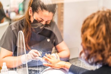 Manikür ve pedikür salonu, koronavirüs, covid-19, sosyal mesafe. Salgın nedeniyle yeniden açılması, güvenlik önlemleri. Koruyucu perdenin arkasında maske takan bir işçinin ayrıntıları