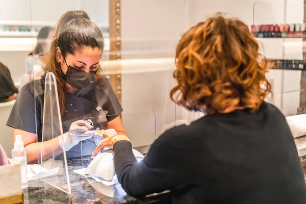 Manicure and pedicure salon, coronavirus, covid-19, social distance. The reopening due to the pandemic, security measures. A female worker behind the protective screen with mask trimmed her nails