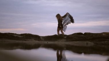 Elbiseyi hareket ettiren kayalıklarda gezinmek. Yazın gün batımında deniz kenarında kayalıklarda şeffaf uzun siyah elbiseli esmer bir kadının yaşam tarzı..