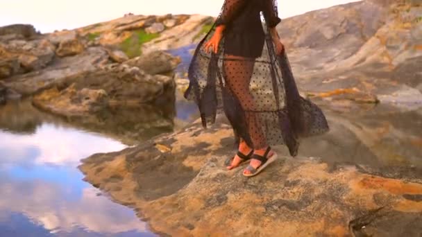 把衣服搬到水中间的岩石上 一个年轻的漂亮的黑发白人女人的生活方式 穿着黑色长裙 在海边夕阳西下 身披透明的衣服 — 图库视频影像