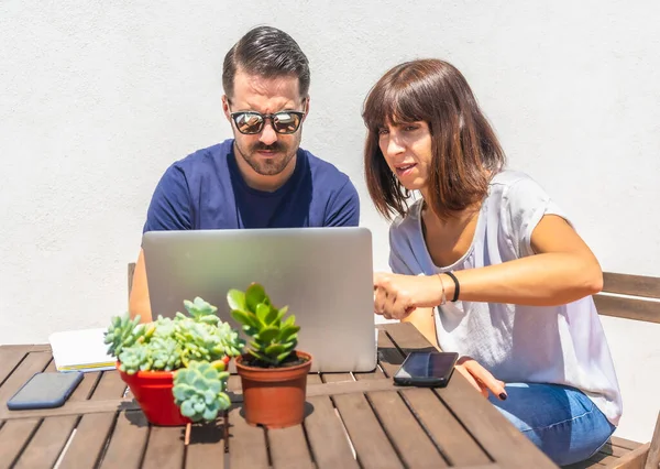 A couple confined at home making a video call with some friends with the computer, on the terrace of their home with a white background sitting at a table. Pointing to an action in the video call