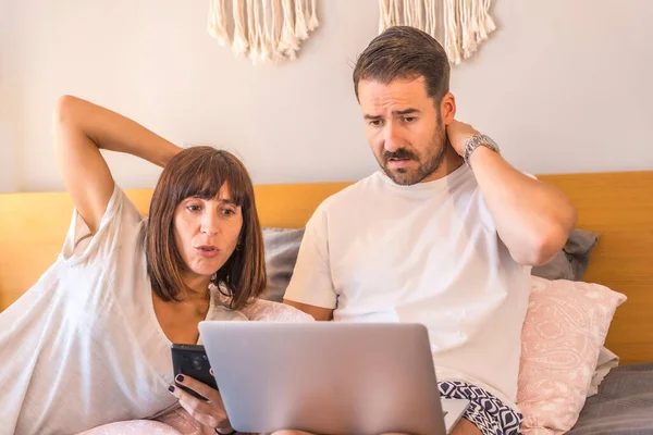 A Caucasian couple on the bed with a computer and a phone, making a reservation at a hotel or flight, organizing vacations, new technologies in family. With doubt of which trip to choose